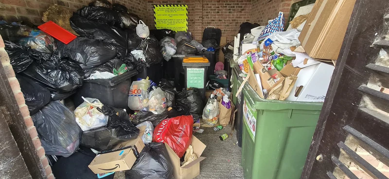 A communal bin store at a block of flats in Gravesend piled to the roof with black rubbish bags, loose boxes and overflowing recycling bins