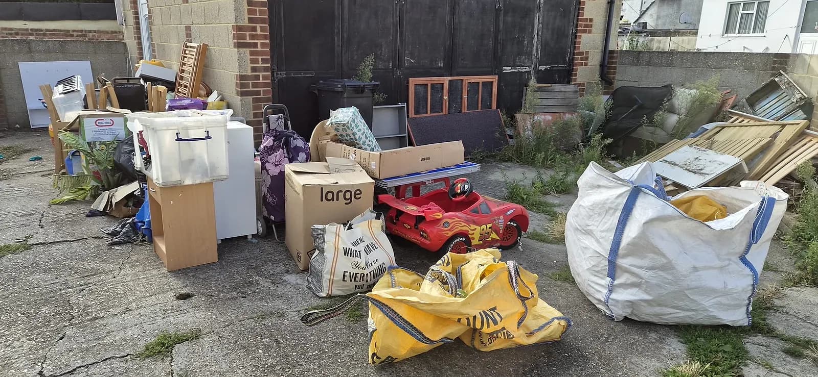 A family's clear-out piled in front of a garage in Gravesend — a kids' ride-on car, chest of drawers, storage crates, a rabbit hutch, boxes and builders' bags