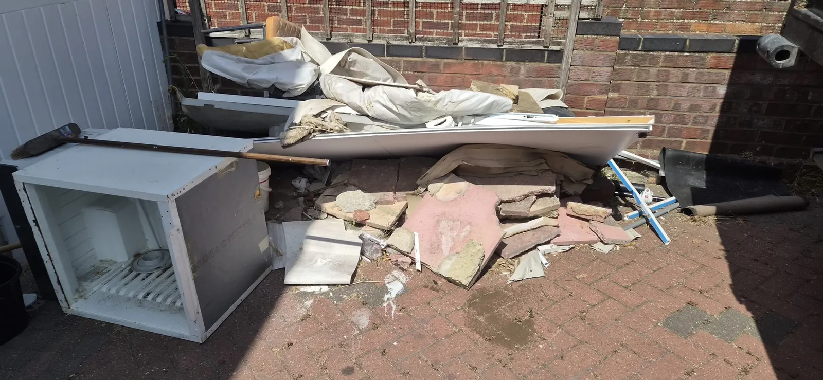An old fridge-freezer on its side with broken slabs, bath panels and dust sheets against a garden wall in Gravesend