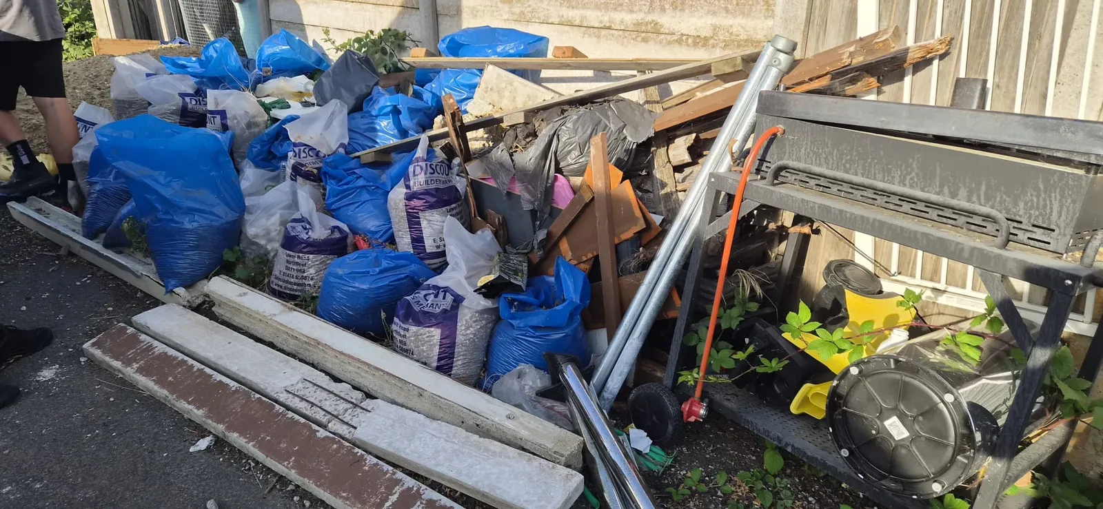 Blue bags of garden waste, builders' sacks and timber stacked along a concrete panel fence in Northfleet