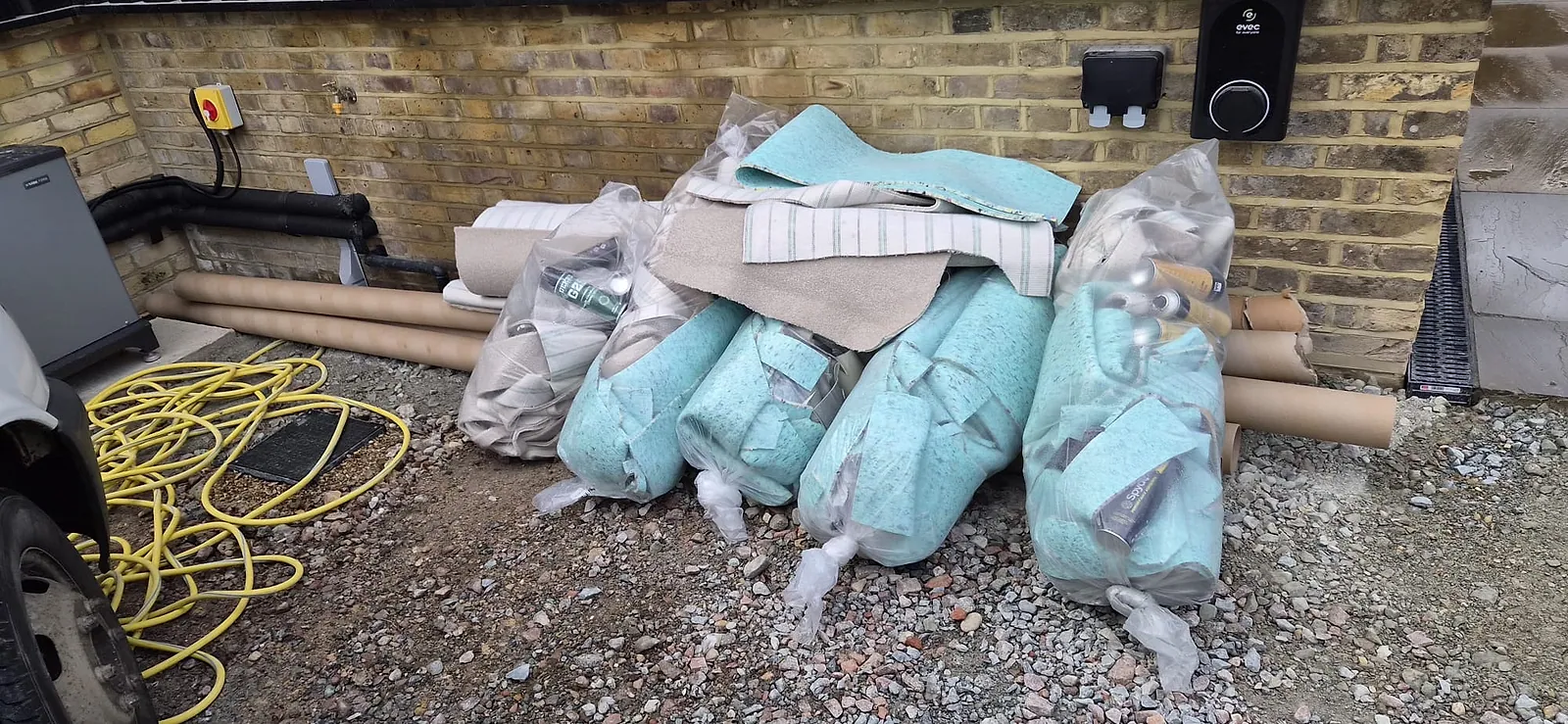 Bales of old carpet underlay in plastic, carpet offcuts and cardboard tubes stacked beside a brick extension in Gravesend