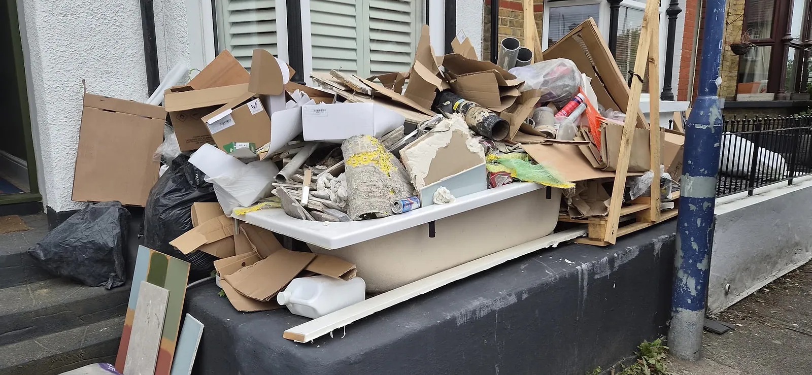 An old bath heaped with plaster, cardboard and pipe offcuts on a front step in Gravesend, pallet and boards stacked beside it