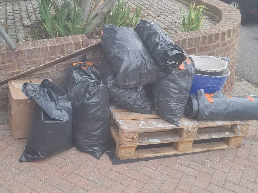Black bags of plaster and rubble stacked on a wooden pallet on a block-paved driveway in Gravesend, with a trug of set plaster